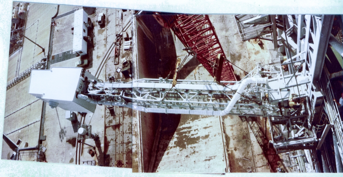 Viewed from farther up on the Fixed Service Structure at Space Shuttle Launch Complex 39-B, to which it is being fastened by union ironworkers from Local 808, the Orbiter Access Arm stands out in sharp relief against the pad deck and flame trench far beneath it, as it hangs still suspended from the crane that lifted it into place, the red boom of which can be seen extending from ground level behind the OAA, and extending up and out of the top of the frame above it. The OAA weighs over 50,000 pounds, and everyone involved is trusting the crane, and the crane operator, with their lives as they continue the work.